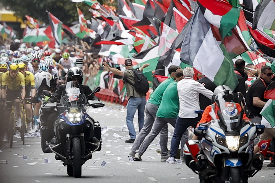 La imagen muestra una escena dinámica de un evento ciclista en una carretera. En primer plano, dos motocicletas avanzan. Detrás, un grupo de ciclistas con maillots de colores y cascos pedalean. A la derecha hay un grupo de manifestantes, entre los que destaca un hombre con los brazos extendidos hacia los ciclistas, en un gesto de protesta (sin tocarles ni acercarse). El fondo de la imagen está dominado por una multitud de banderas ondeando densamente. Se distinguen claramente numerosas banderas palestinas y algunas vascas.