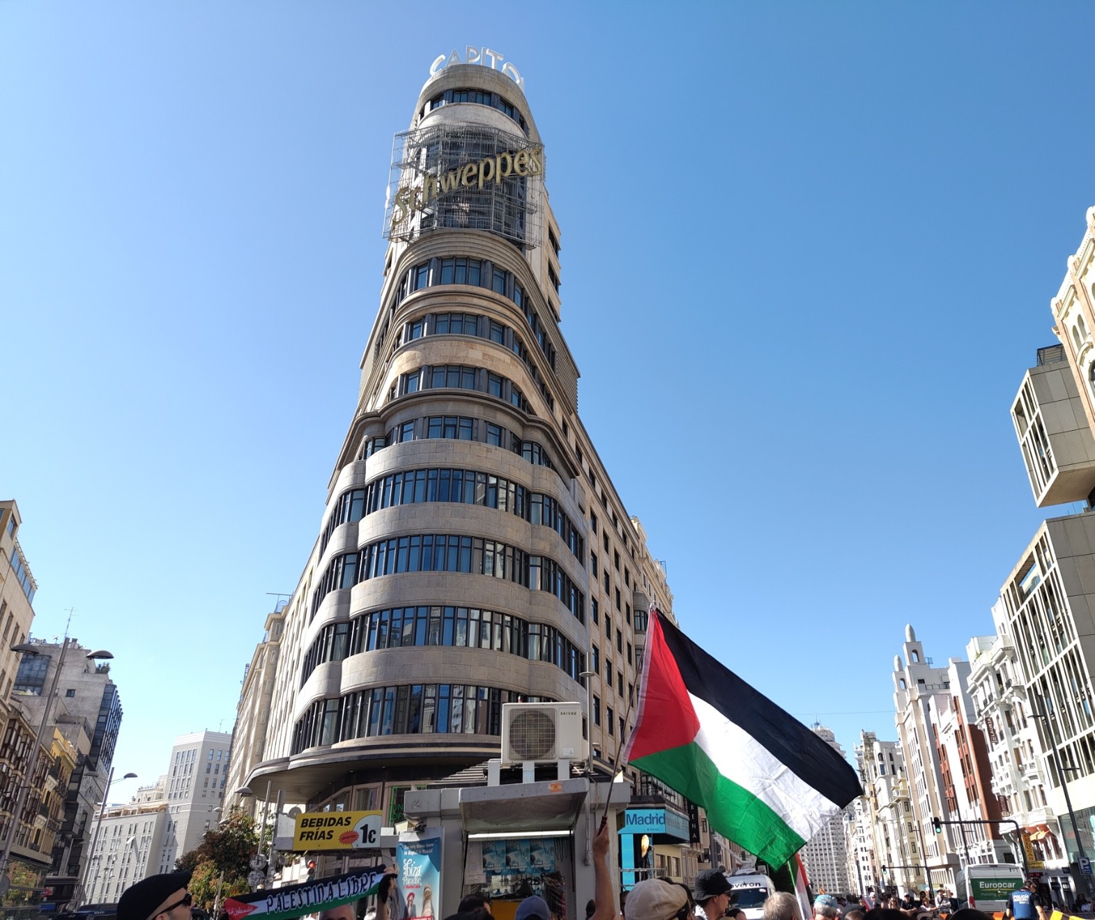 Fotografía del icónico edificio con un cartel publicitario de Schweppes que está situado en la plaza de Callao, en Madrid. Delante, ondea una bandera de Palestina (sujetada por personas que se encuentran debajo, no aparecen en la foto).