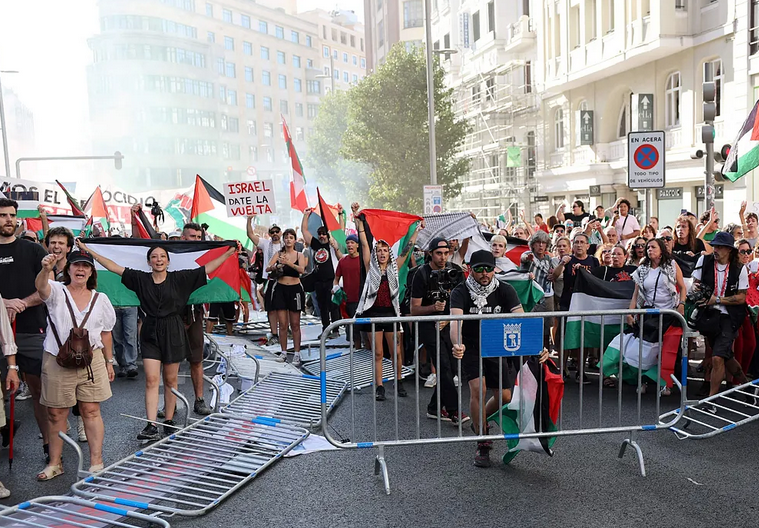 Una vista general de la Gran Vía de Madrid. Numerosos manifestantes, de diversas edades, llenan la calle. La mayoría porta banderas palestinas y algunos llevan keffiyehs. Se pueden ver pancartas con mensajes como "ISRAEL DATE LA VUELTA" y "PAREMOS EL GENOCIDIO". En primer plano, varias vallas metálicas de seguridad están tumbadas en el suelo y otras siendo empujadas por los manifestantes. Una de las vallas tiene el escudo del Ayuntamiento de Madrid. Les participantes muestran expresiones de determinación, con algunos puños levantados.